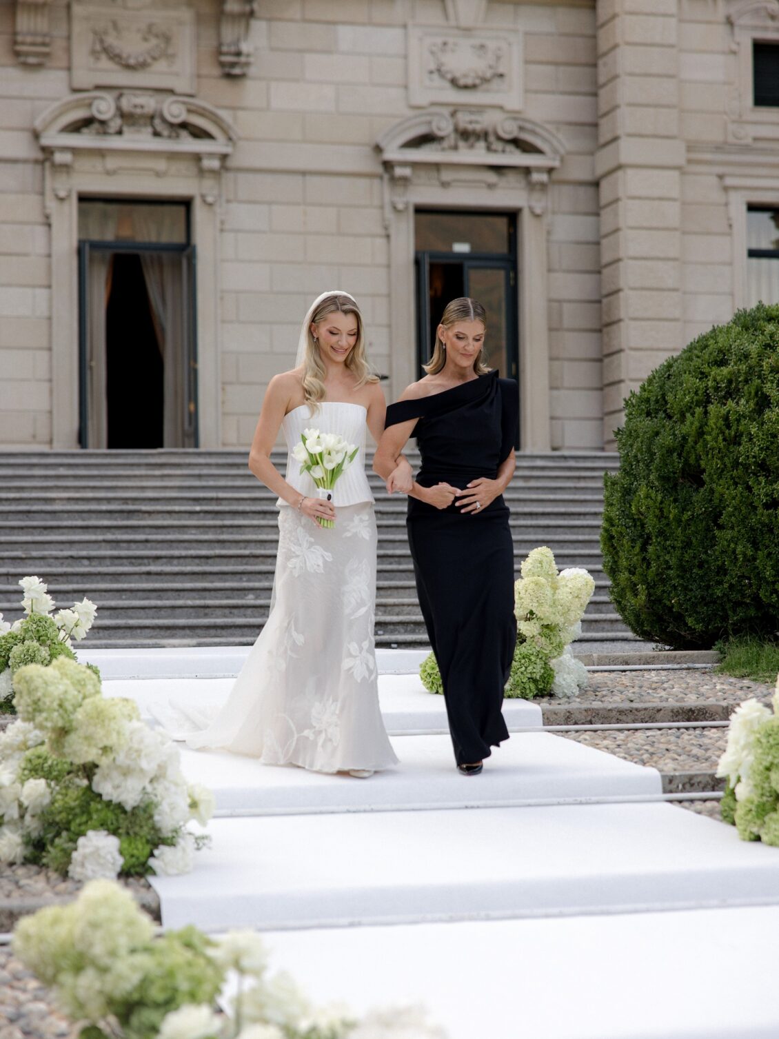 Bride walks down the aisle arm in arm with a woman in black.