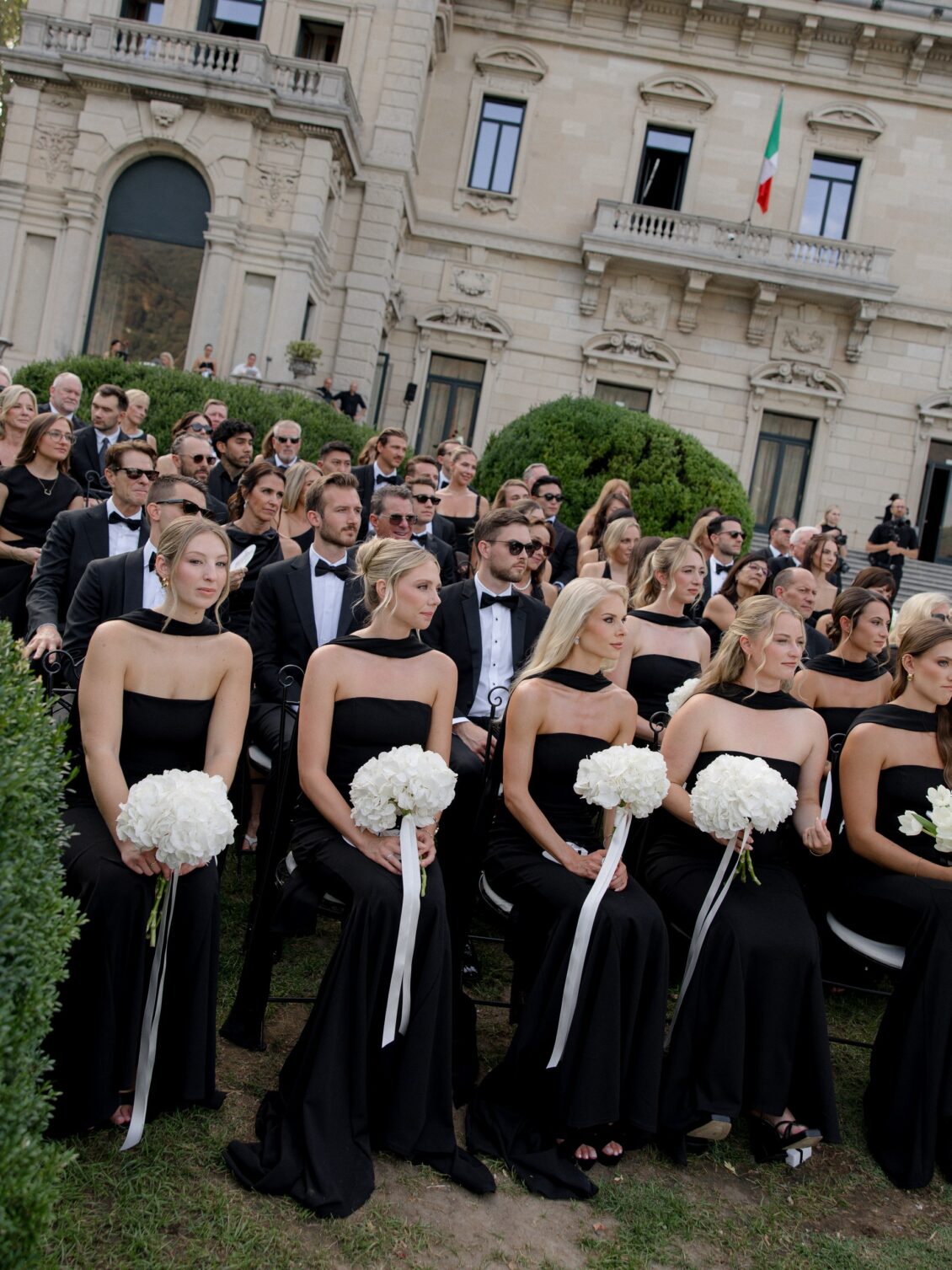 Attendants in black gowns hold white bouquets before seated guests.