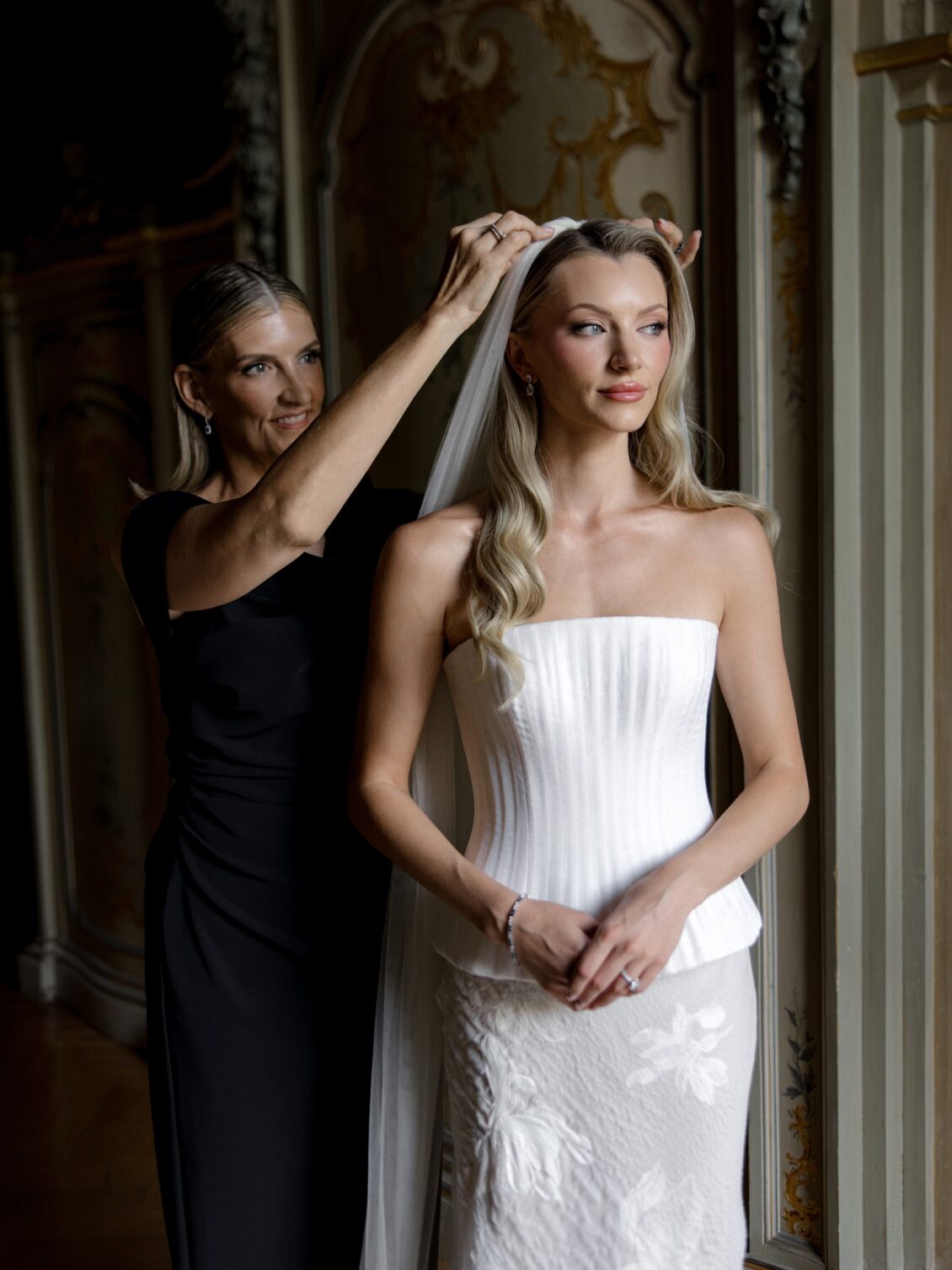 Attendant adjusts the bride’s hair beside a window.