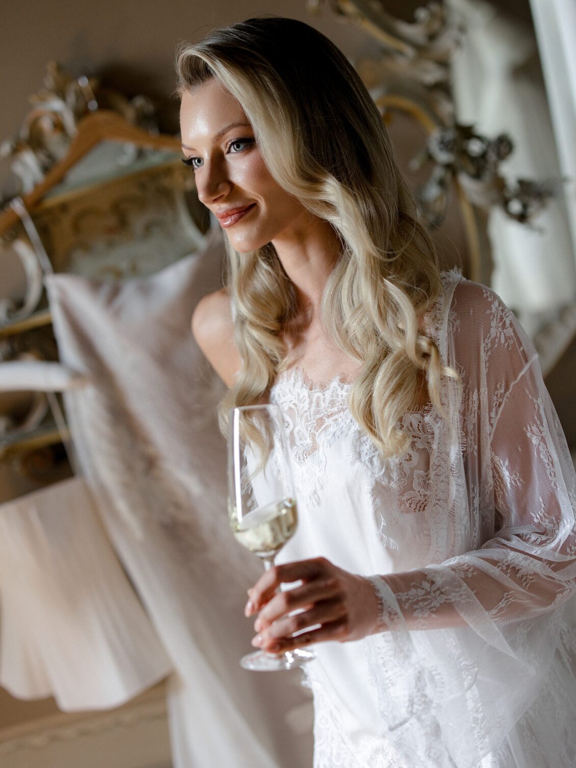 Bride in a lace robe holds a champagne glass beside an ornate mirror.