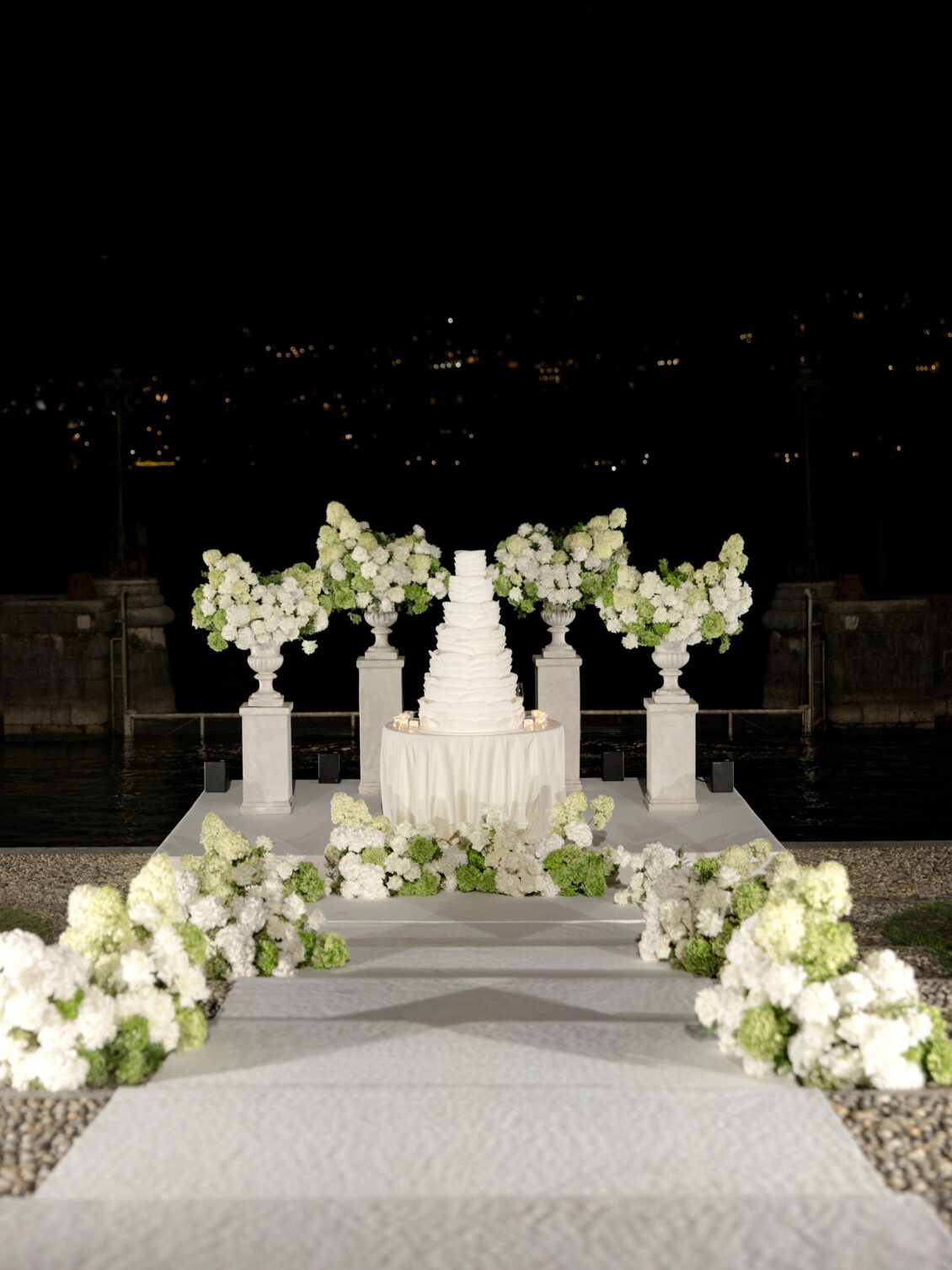 Tiered white wedding cake framed by white flowers at night.