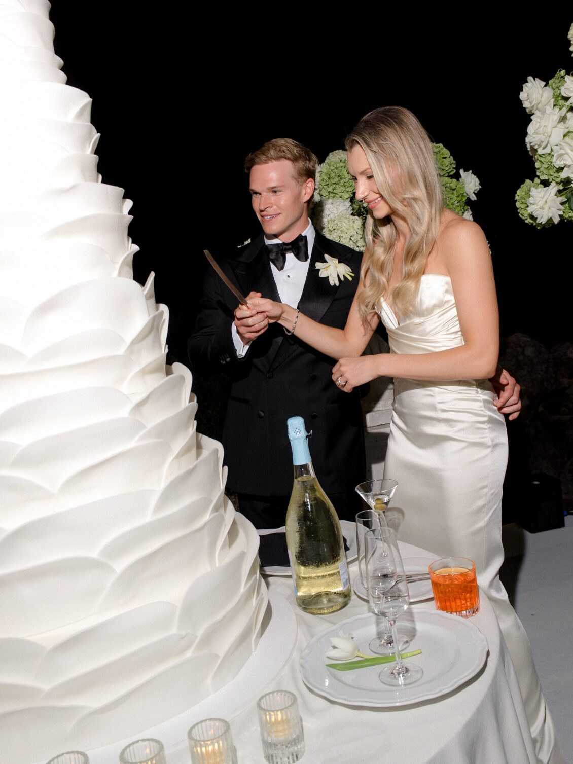 Newlyweds cut their tall white wedding cake outdoors at night.