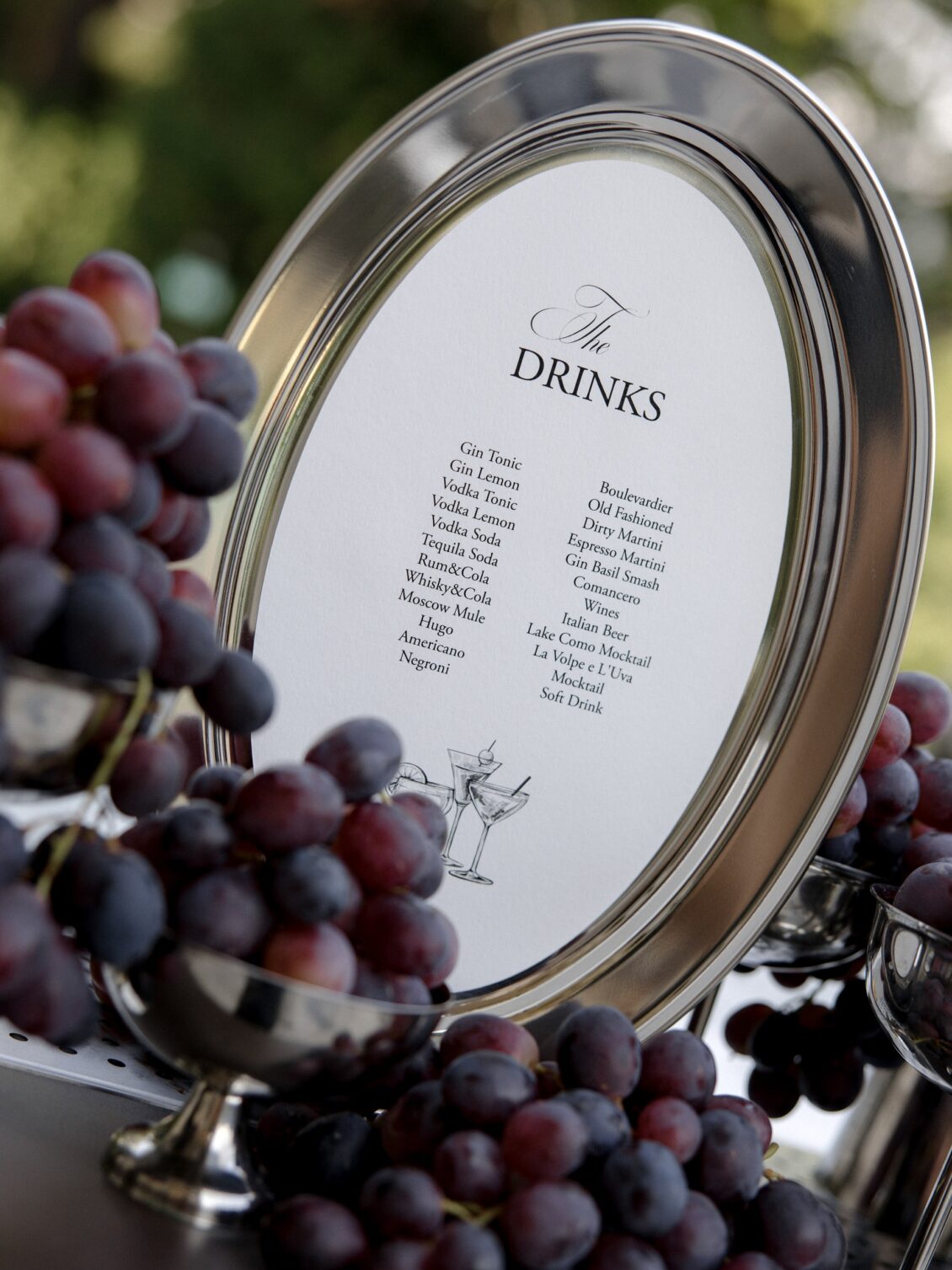 Silver-framed drinks menu displayed among purple grapes.