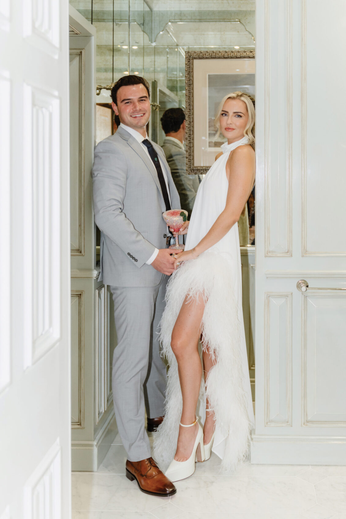 A man in a light gray suit and a woman in a white feathered dress stand together indoors, holding hands and smiling, with elegant white paneling and a mirror behind them—evoking the timeless romance of an Irish castle wedding at Ashford Castle.