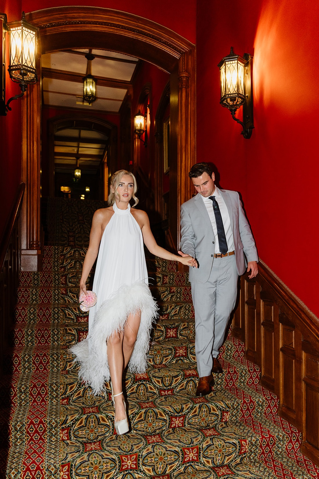A woman in a white feathered dress and a man in a light gray suit walk down a patterned carpeted staircase in a building with red walls and ornate lighting, capturing the elegance of an Irish castle wedding at Ashford Castle.
