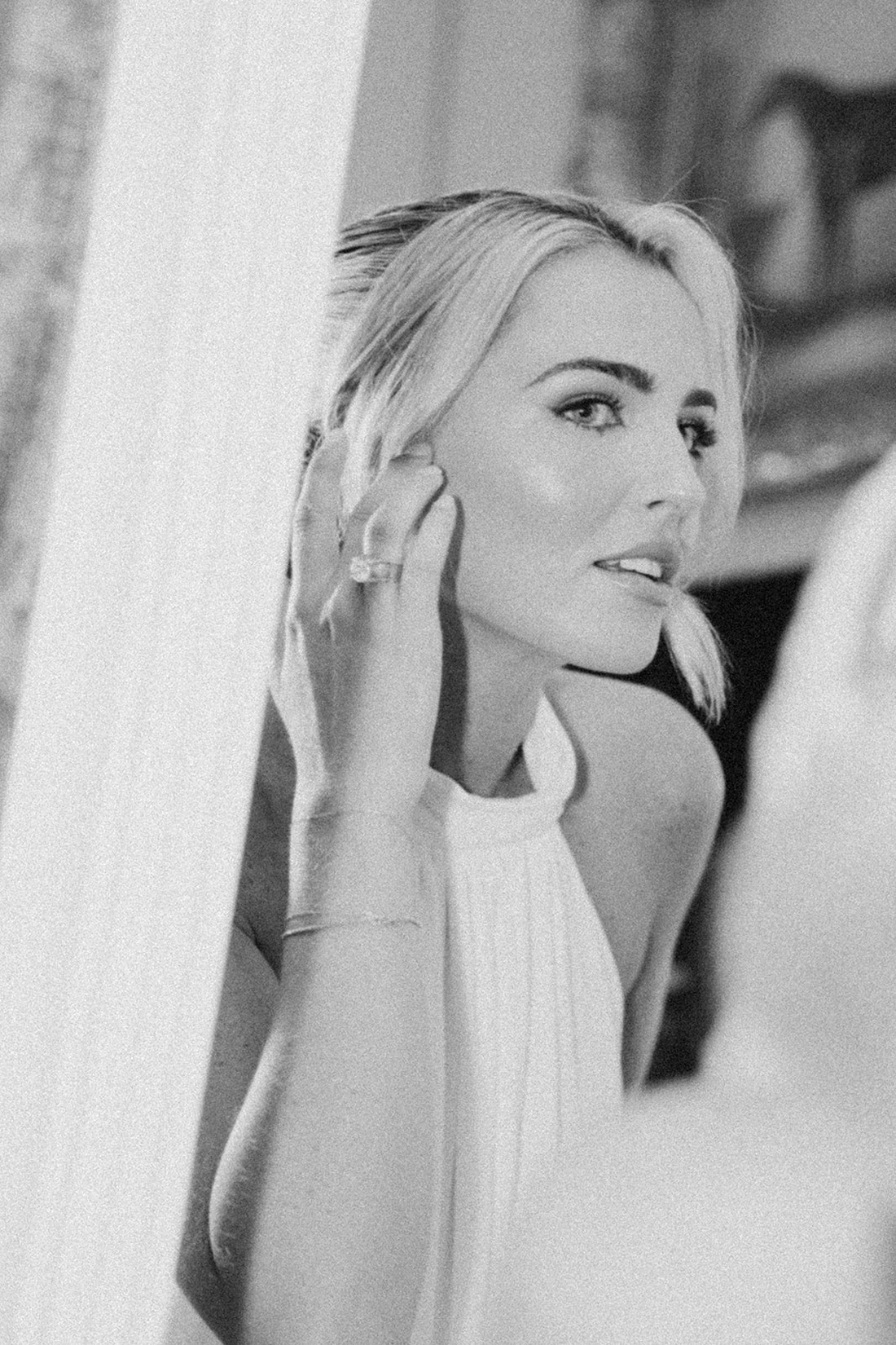 A woman in a white halter dress looks at herself in a mirror, touching her ear. The photo is in black and white, highlighting her elegant pose and soft features as she prepares for an Irish castle wedding at Ashford Castle.