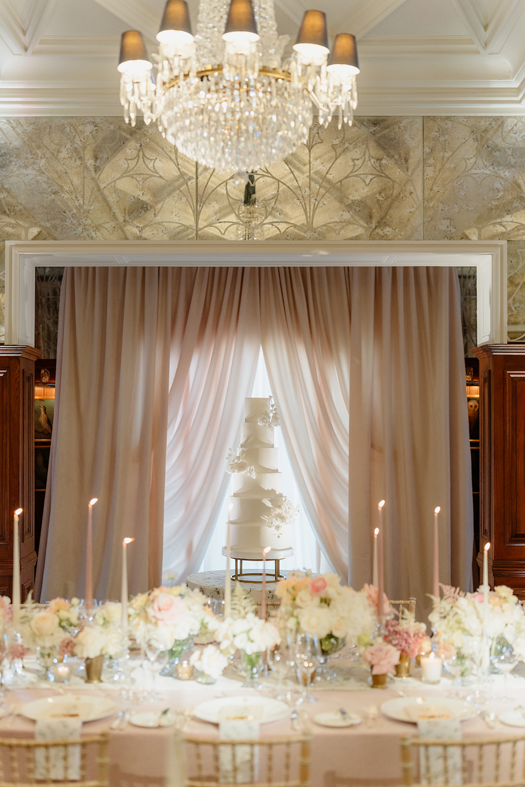 A tall white wedding cake with floral decorations stands in front of sheer pink curtains, surrounded by elegant table settings with flowers, candles, and glassware under a crystal chandelier—capturing the timeless romance of an Irish castle wedding at Ashford Castle.