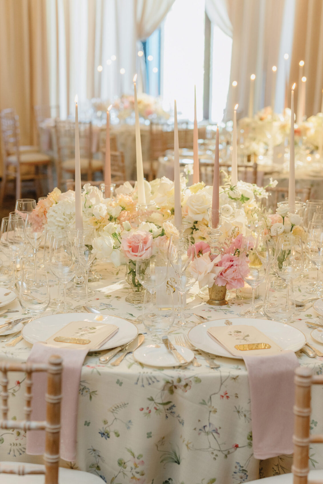 Elegant table setting with pastel flowers, tall taper candles, glassware, and gold-accented place cards on a white floral tablecloth, with wooden chairs and light pink napkins. The soft, romantic ambiance evokes the timeless charm of an Irish castle wedding at Ashford Castle.