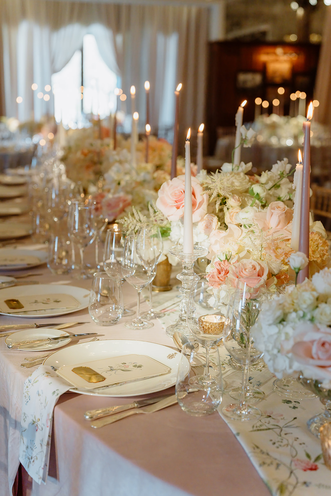Elegant table set for a formal event with floral arrangements, tall candle holders, glassware, plates, and napkins on a pastel tablecloth—perfect for an Irish castle wedding at Ashford Castle—surrounded by soft lighting and draped curtains in the background.