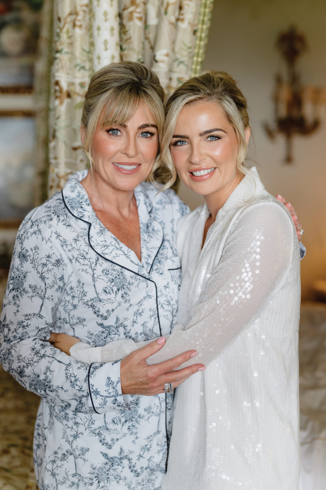 Two women stand close together, smiling and embracing. One wears a floral pajama top; the other wears a white, sparkly robe. They appear happy in a warmly lit, elegant room, the atmosphere capturing the joy of an Irish castle wedding at Ashford Castle.