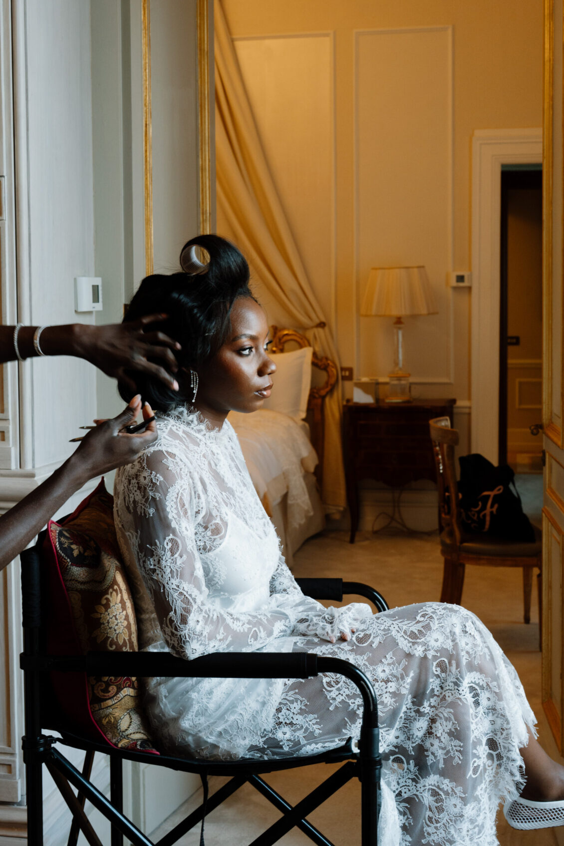 A woman in a white lace dress sits on a chair, having her hair styled in a softly lit, elegant room with a bed and lamp in the background, preparing for her Nigerian wedding in Rome.