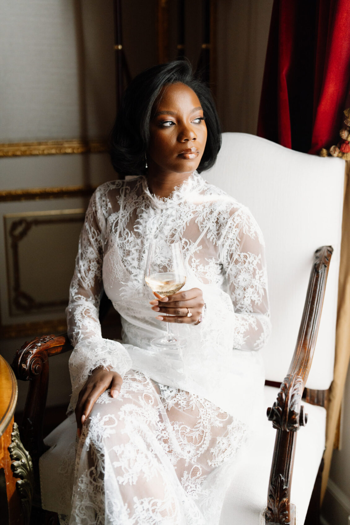 A woman in a white lace dress sits in an elegant chair, holding a glass of wine and looking thoughtfully to the side, capturing the refined atmosphere of a Nigerian wedding in Rome.