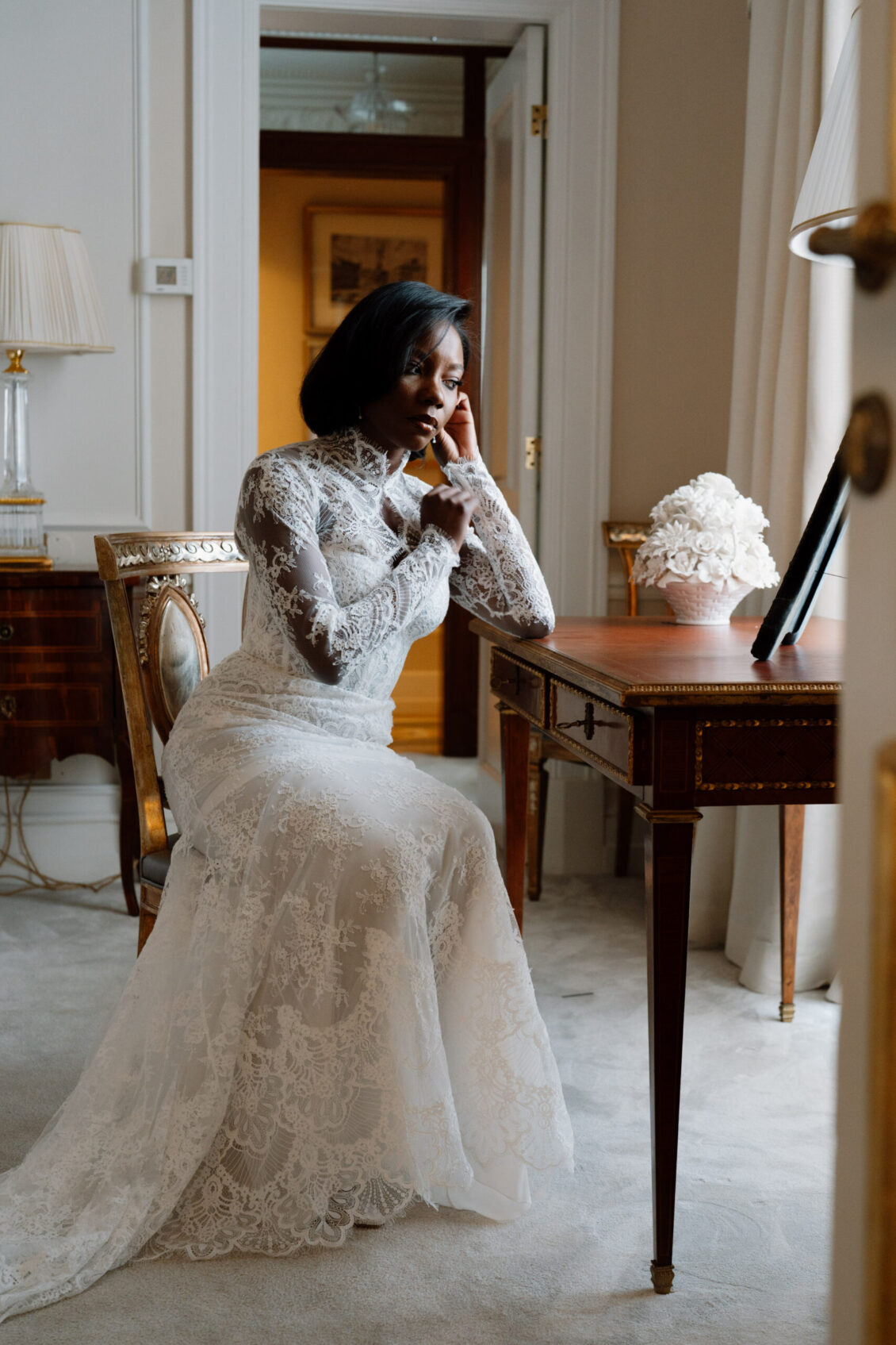 A woman in a long-sleeved lace wedding dress sits pensively at an ornate wooden desk in a softly lit, elegant room, capturing the serene anticipation of a Nigerian wedding in Rome.