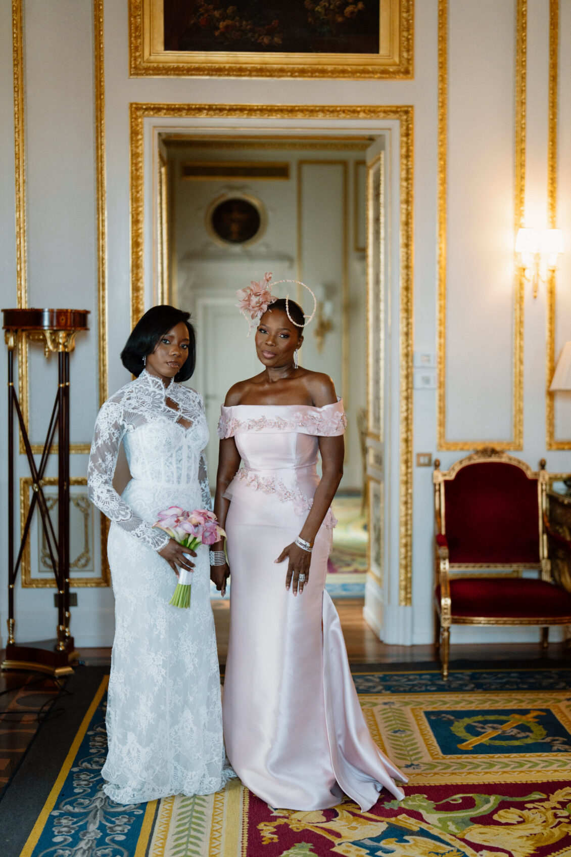 Two elegantly dressed women stand side by side in a lavish, gold-trimmed room at a Nigerian wedding in Rome. One wears a white lace gown and holds pink flowers; the other wears a pale pink off-shoulder dress with a floral headpiece.