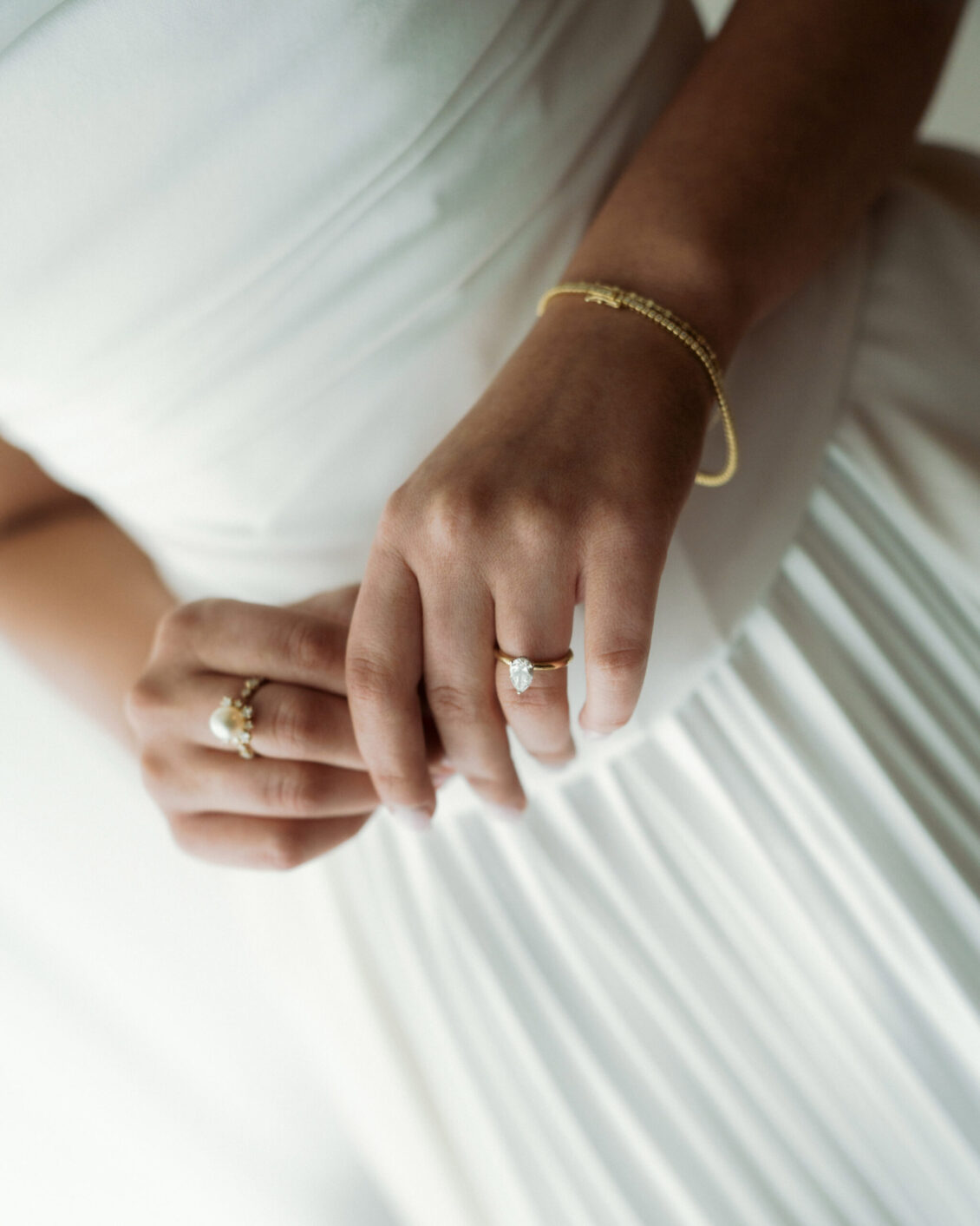 A person in a white dress shows their hands, adorned with gold rings and a delicate bracelet, highlighting an engagement ring with a large gemstone—an image reminiscent of the timeless elegance seen at the Ariana and Julian Borghese wedding.