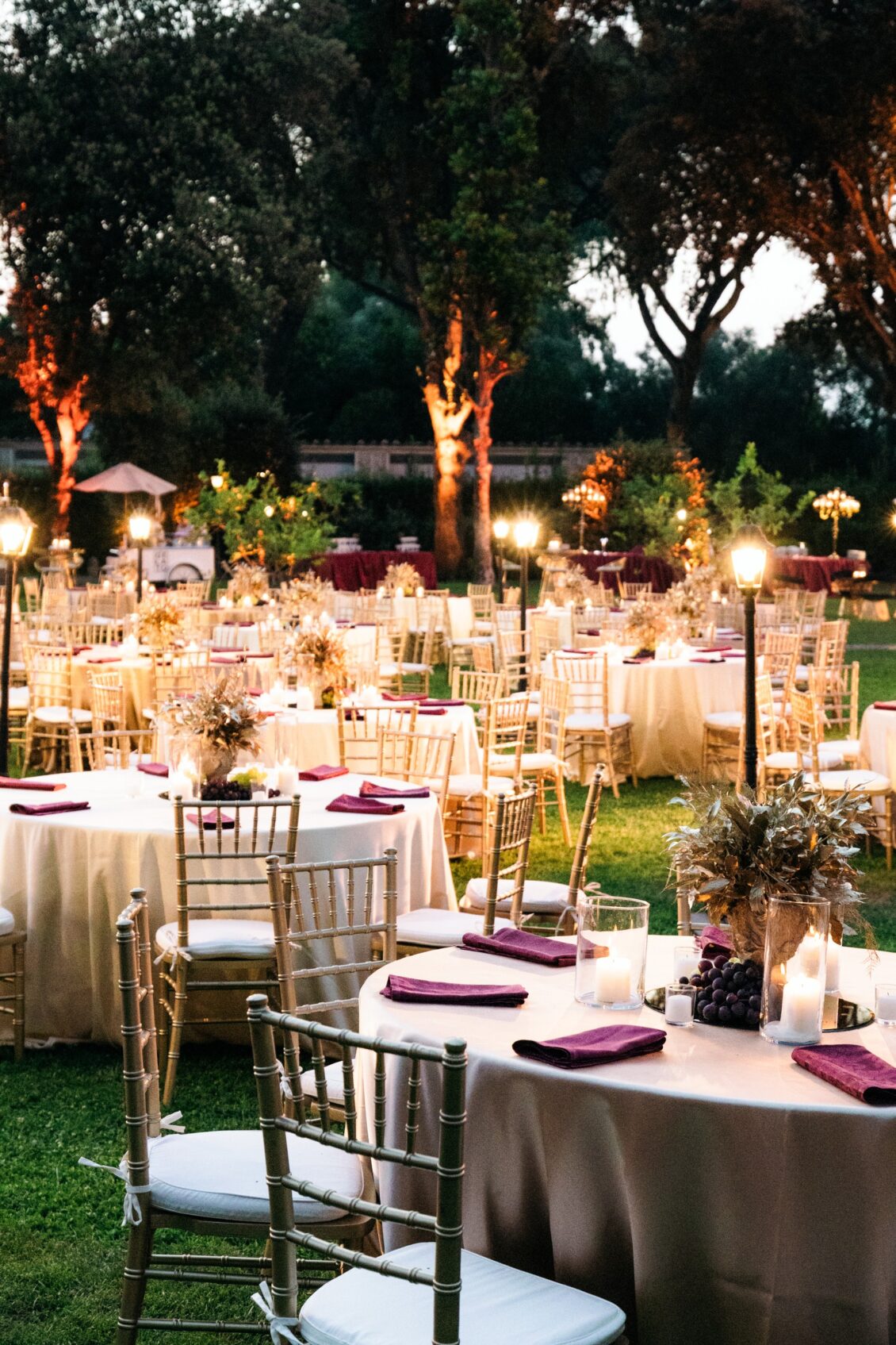 Gold chairs and candlelit round tables with purple napkins beneath illuminated trees at a garden dinner in Rome.