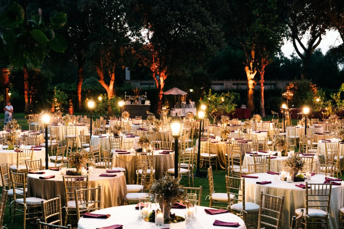 Wide view of an empty candlelit garden reception with round tables, gold chairs and illuminated trees in Rome.