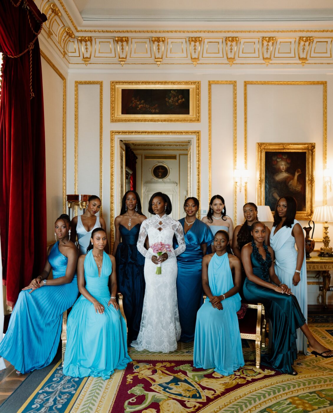 Bride in a white lace dress with eleven attendants in blue gowns posing in an ornate gilded room.