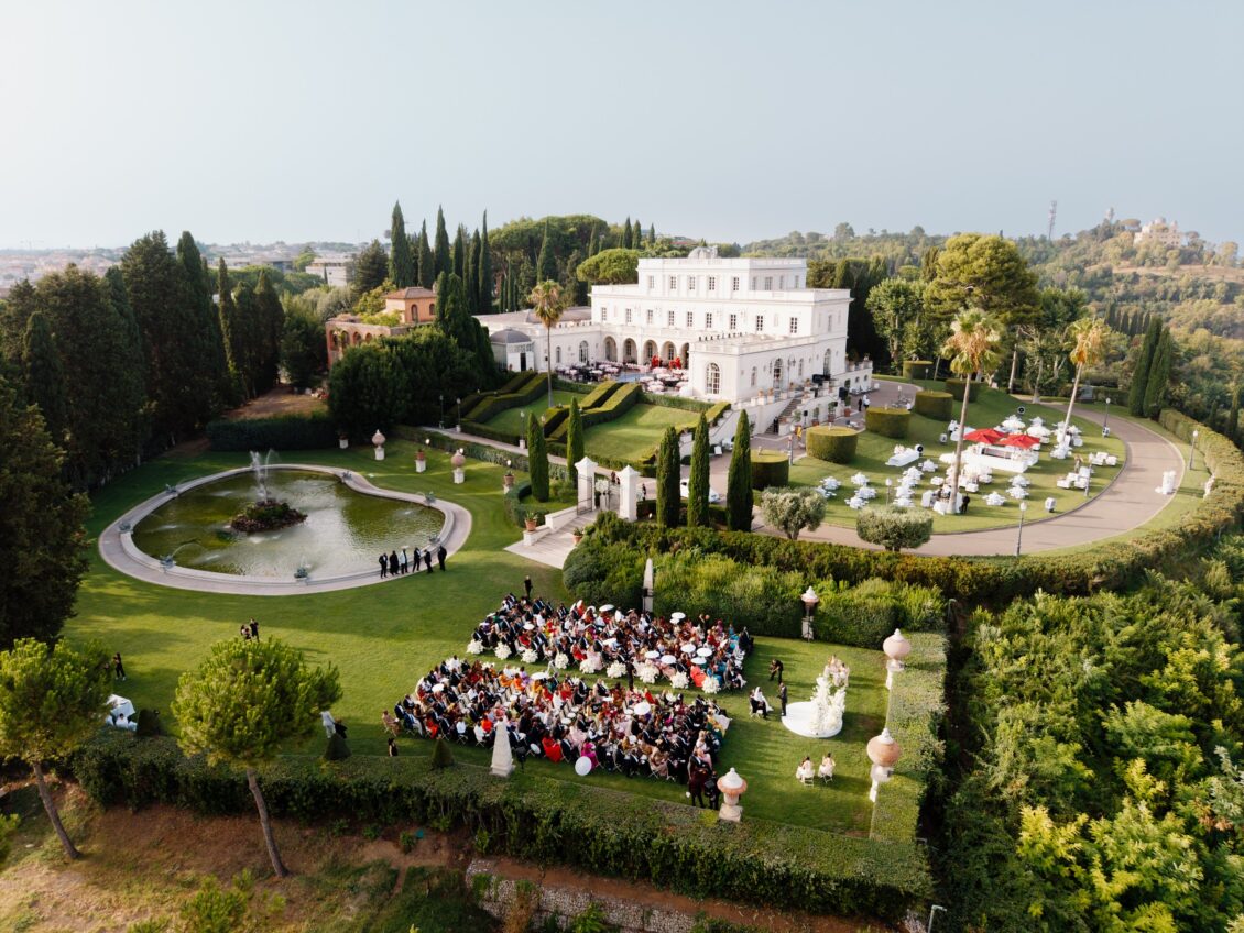 Aerial view of guests seated for an outdoor wedding ceremony beside a fountain and white villa in Rome.