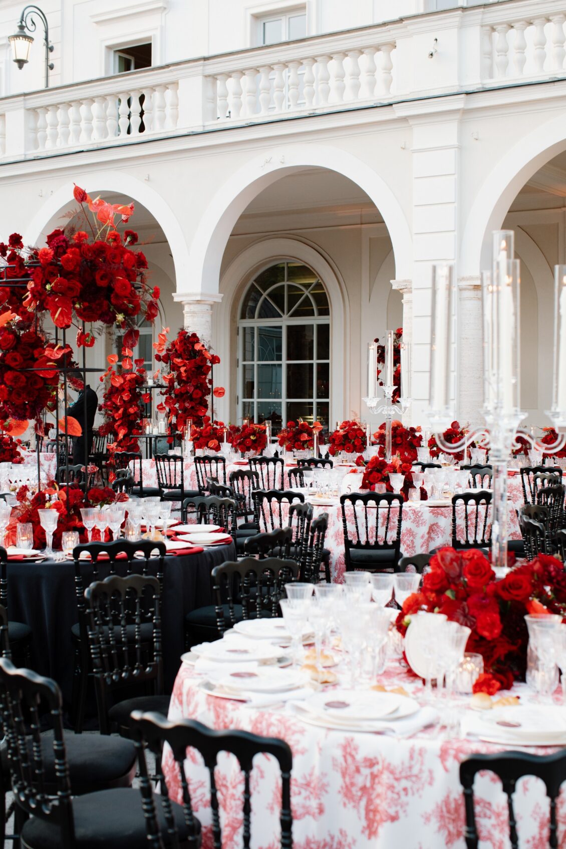 Terrace tables with black chairs, patterned linens, tall candles and red flowers beside the white arches of a Rome villa.
