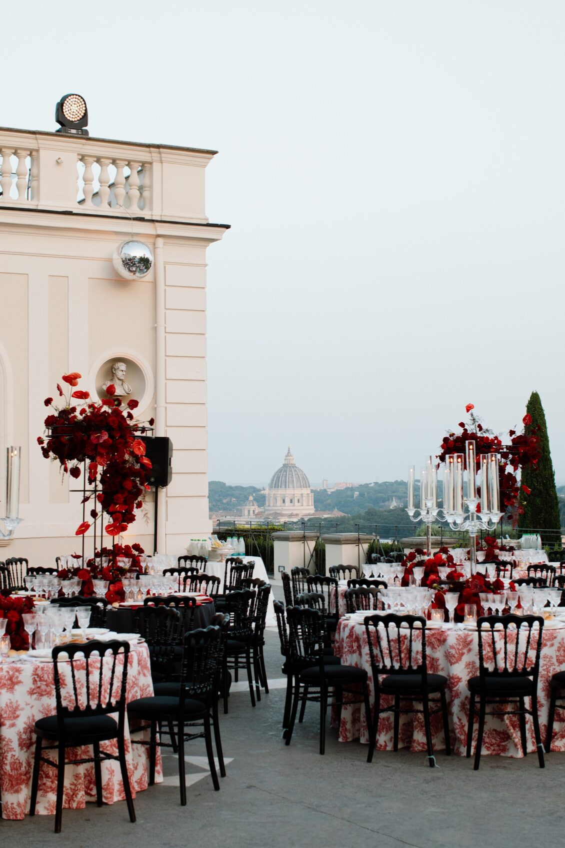 Red-and-white wedding tables on a villa terrace with black chairs and a domed Rome skyline in the distance.