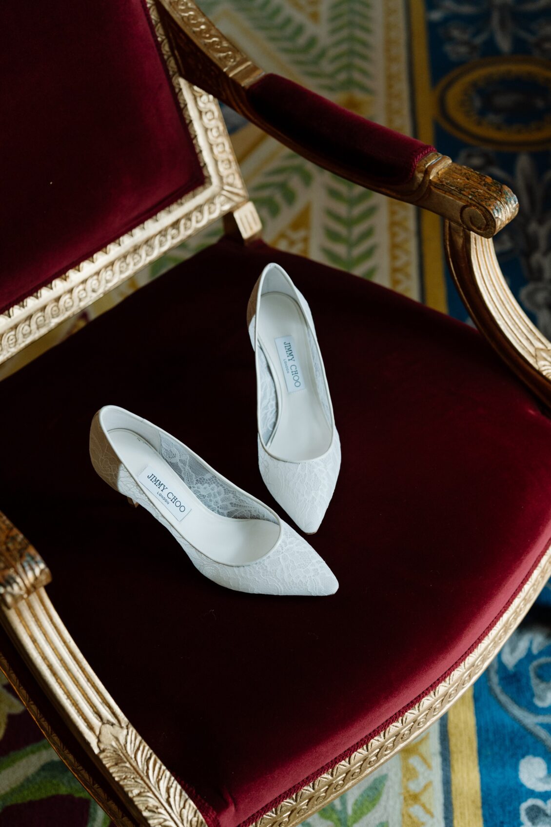 White lace Jimmy Choo bridal pumps displayed on a burgundy velvet and gold antique chair.