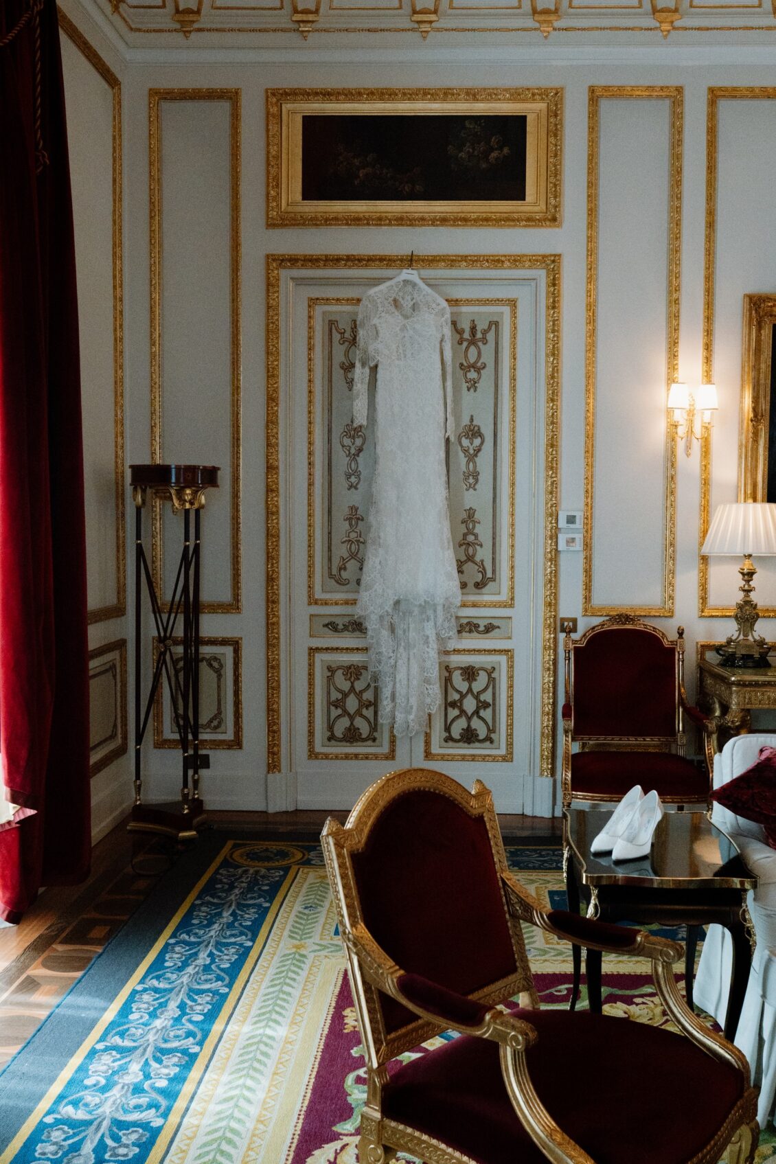 White lace wedding dress hanging on a decorated door in an ornate cream-and-gold room with burgundy chairs.