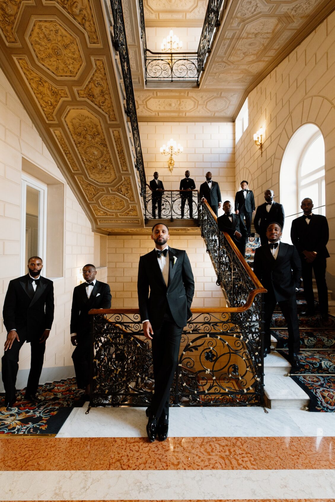 Groom and nine groomsmen in black tuxedos posing across an ornate marble staircase in Rome.