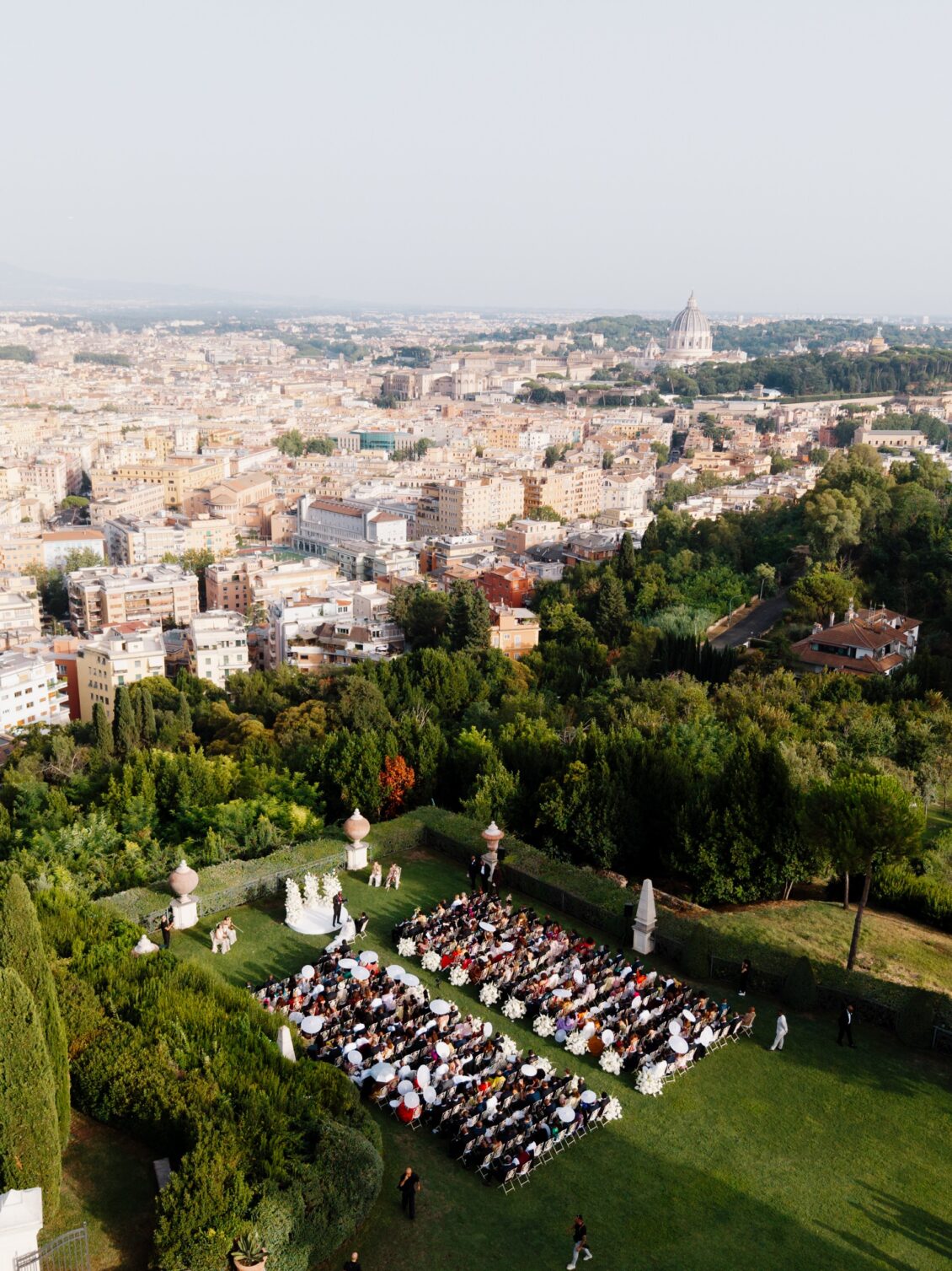 Guests attend an outdoor wedding ceremony on a formal villa lawn with gardens, the Rome skyline, and a distant dome beyond.