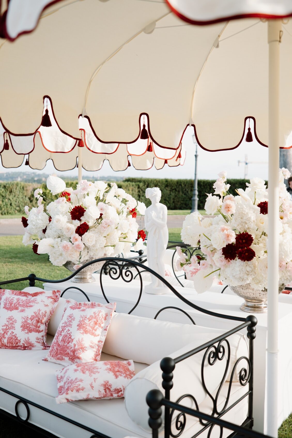 Wrought-iron daybed with patterned cushions beneath cream parasols and large white, blush, and dark-red wedding flowers.