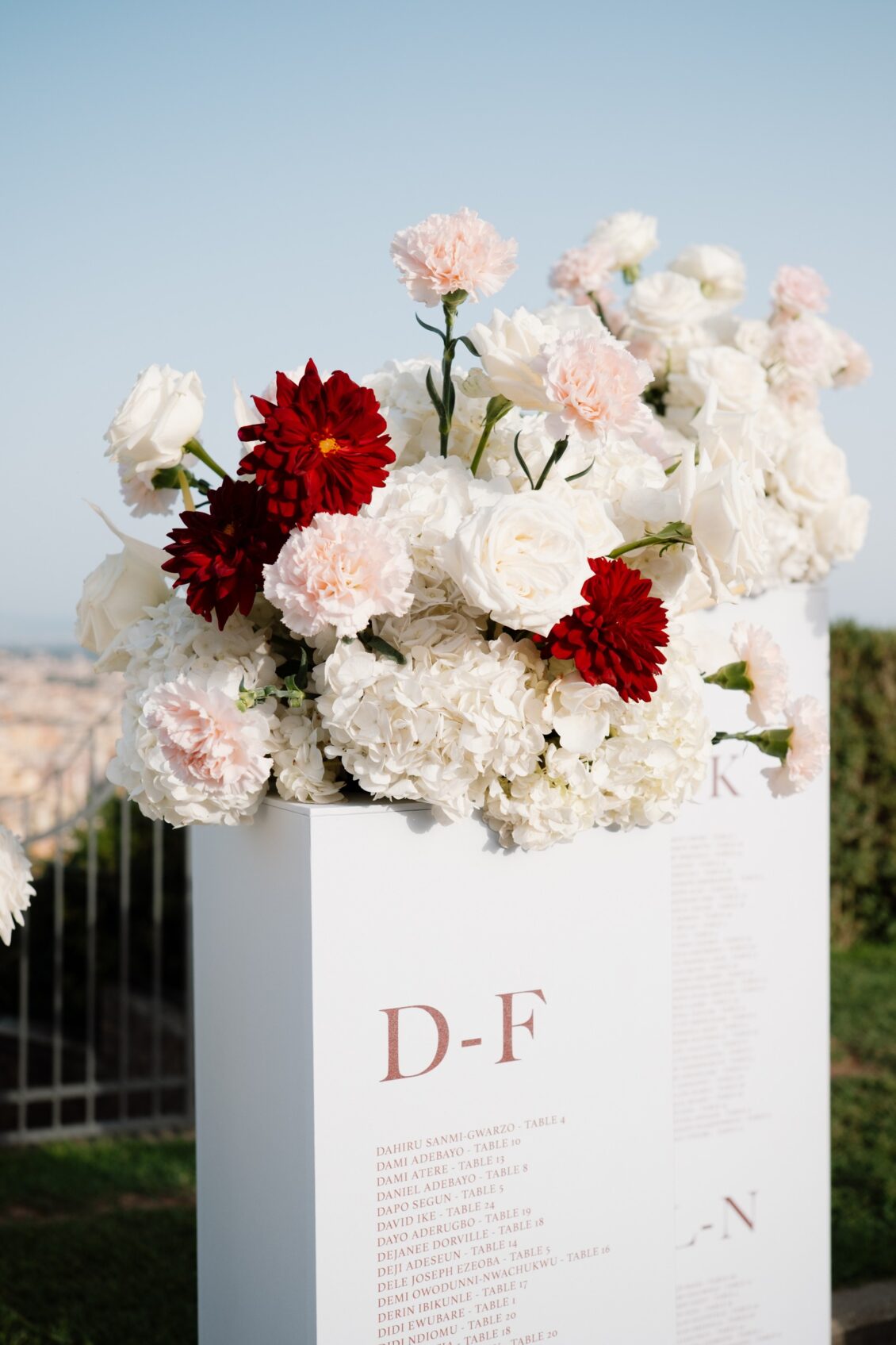 White D–F seating chart topped with white, blush, and dark-red flowers stands outdoors with a soft city view behind it.