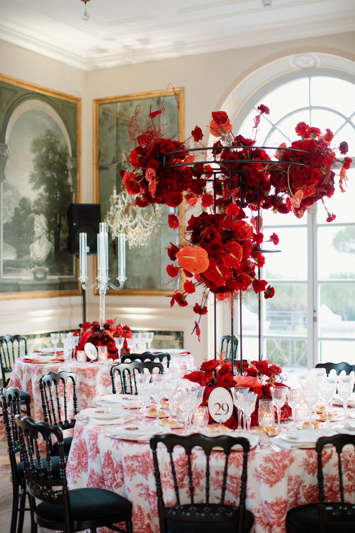 Table 29 in a pale mural-lined villa room with red flowers on a tall black frame, printed linens, black chairs, glassware, and candles.