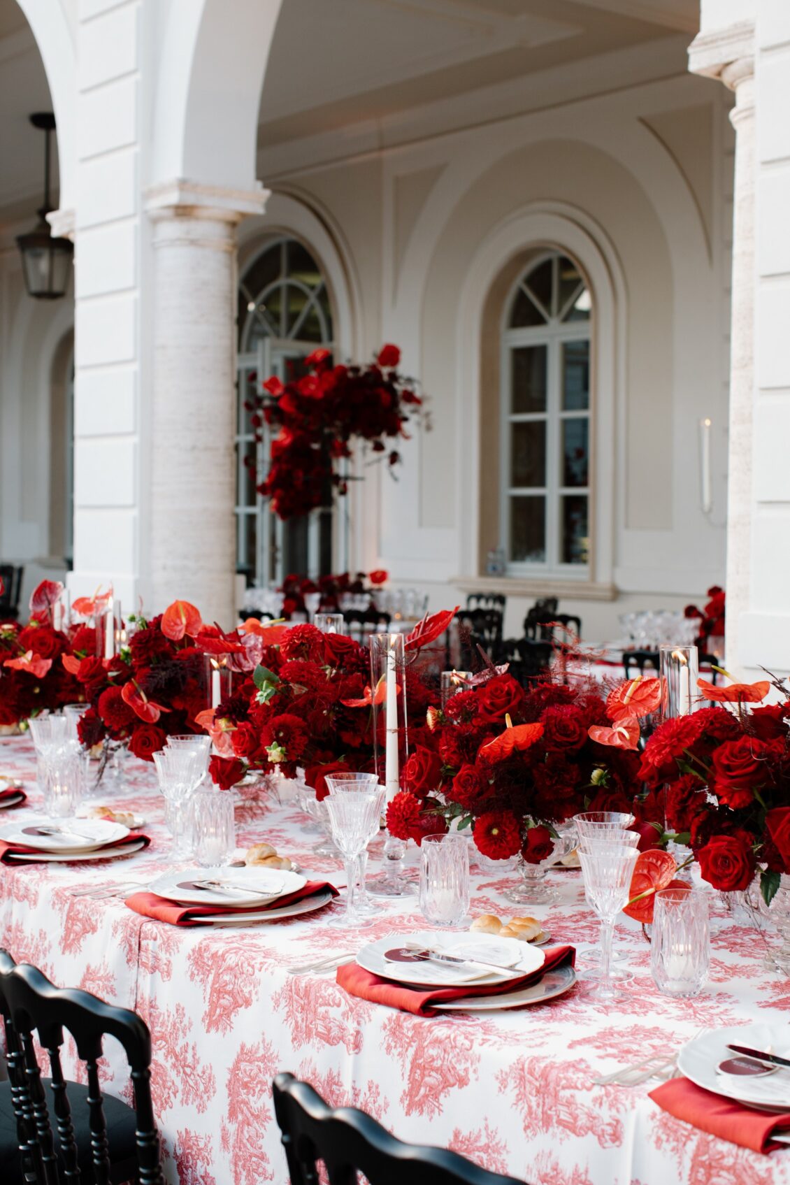 Long villa reception table with red flowers, white plates, red napkins, crystal glasses, candles, bread rolls, and a red-and-white printed cloth.