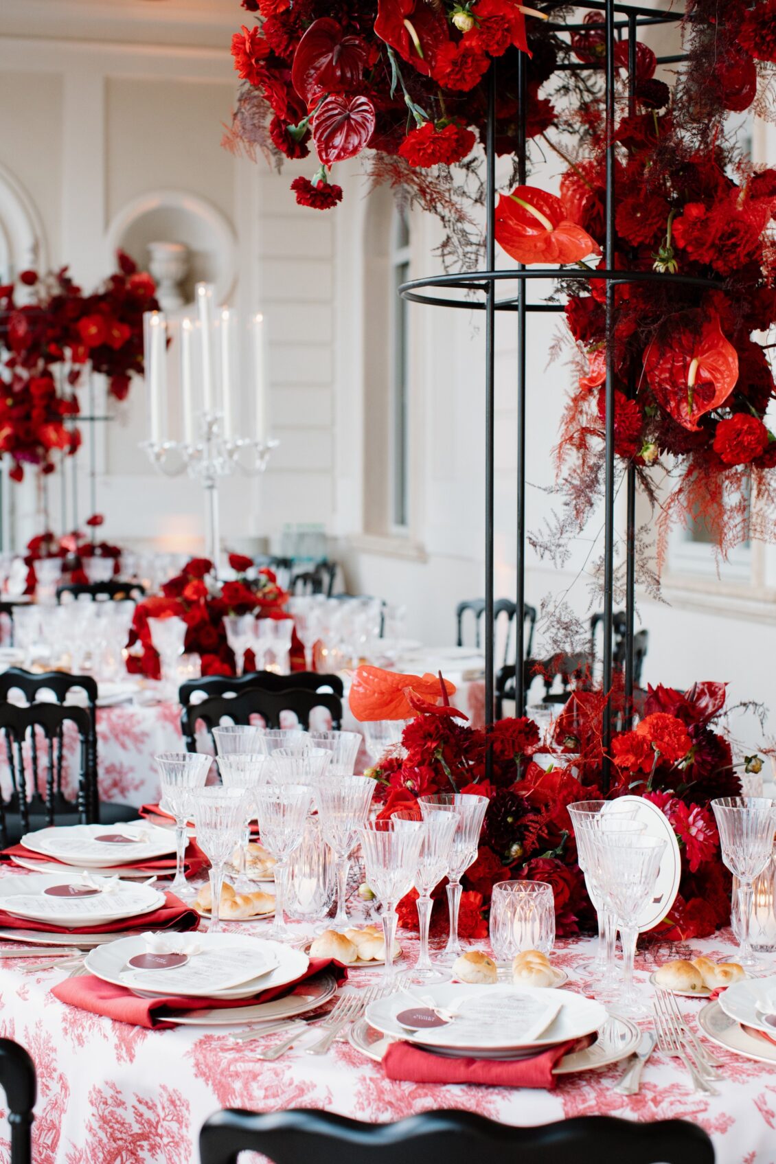 Tall black reception centerpiece filled with red roses, dahlias, anthuriums, and foliage above a table set with crystal glassware and red napkins.