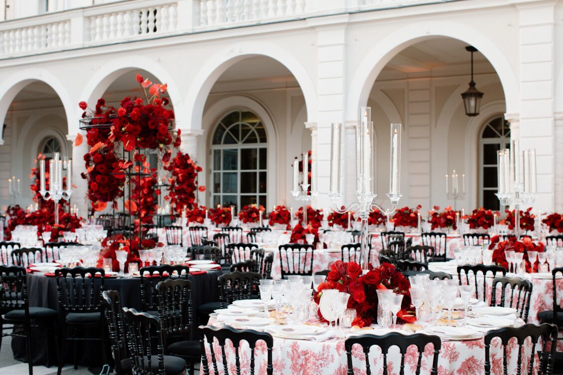 Wide reception terrace filled with black chairs, red flowers, printed tablecloths, and tall candles beneath a white arched Rome villa facade.