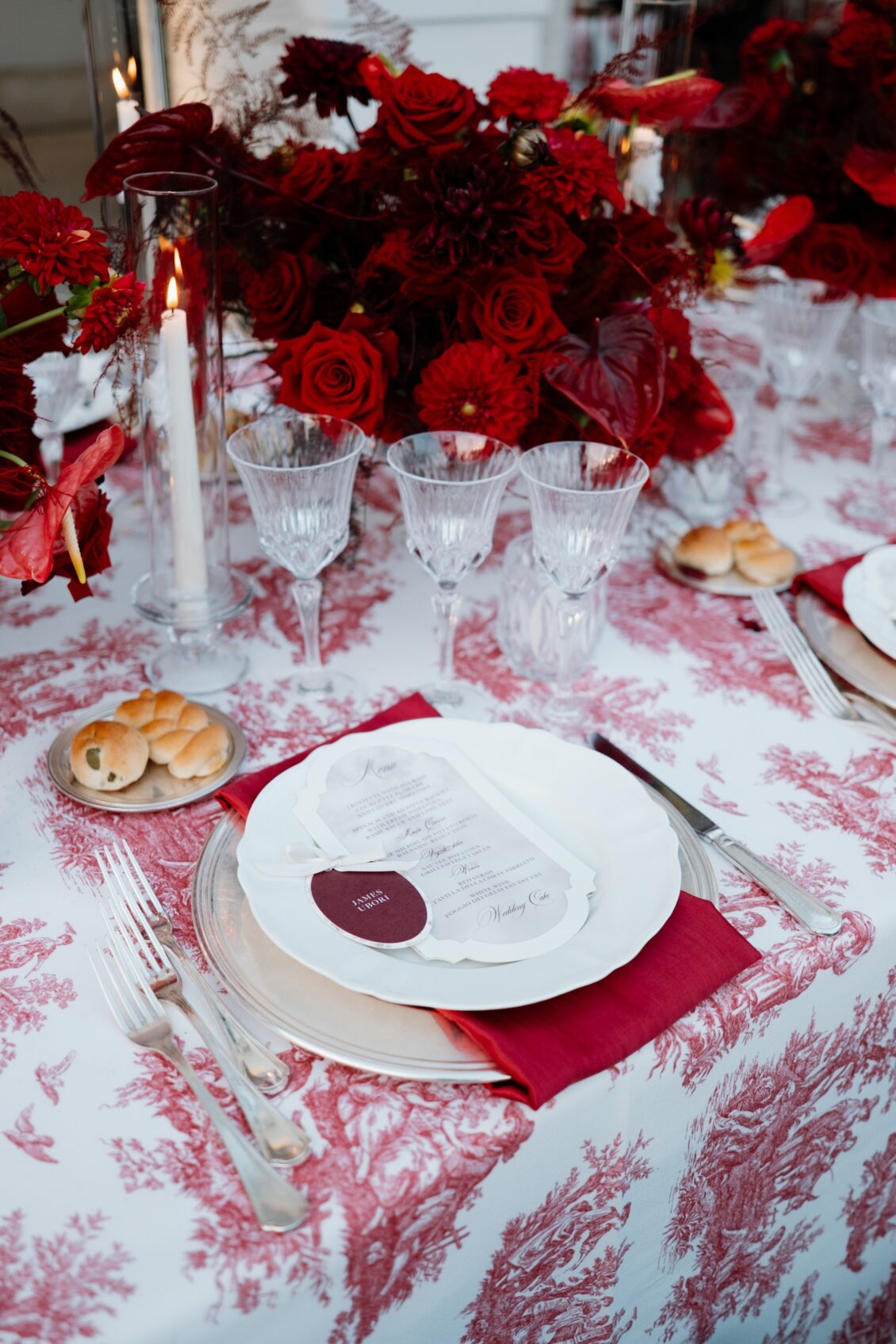 Wedding place setting with clear chargers, white plates, red napkin, menu, burgundy name card, crystal glasses, bread, candles, and red flowers.