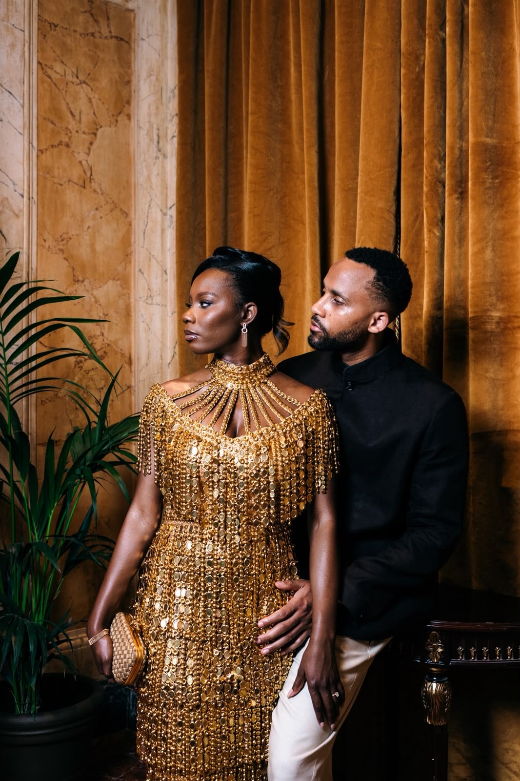 Couple posing beside amber stone columns, with the woman in a gold beaded gown and the man in a black jacket.