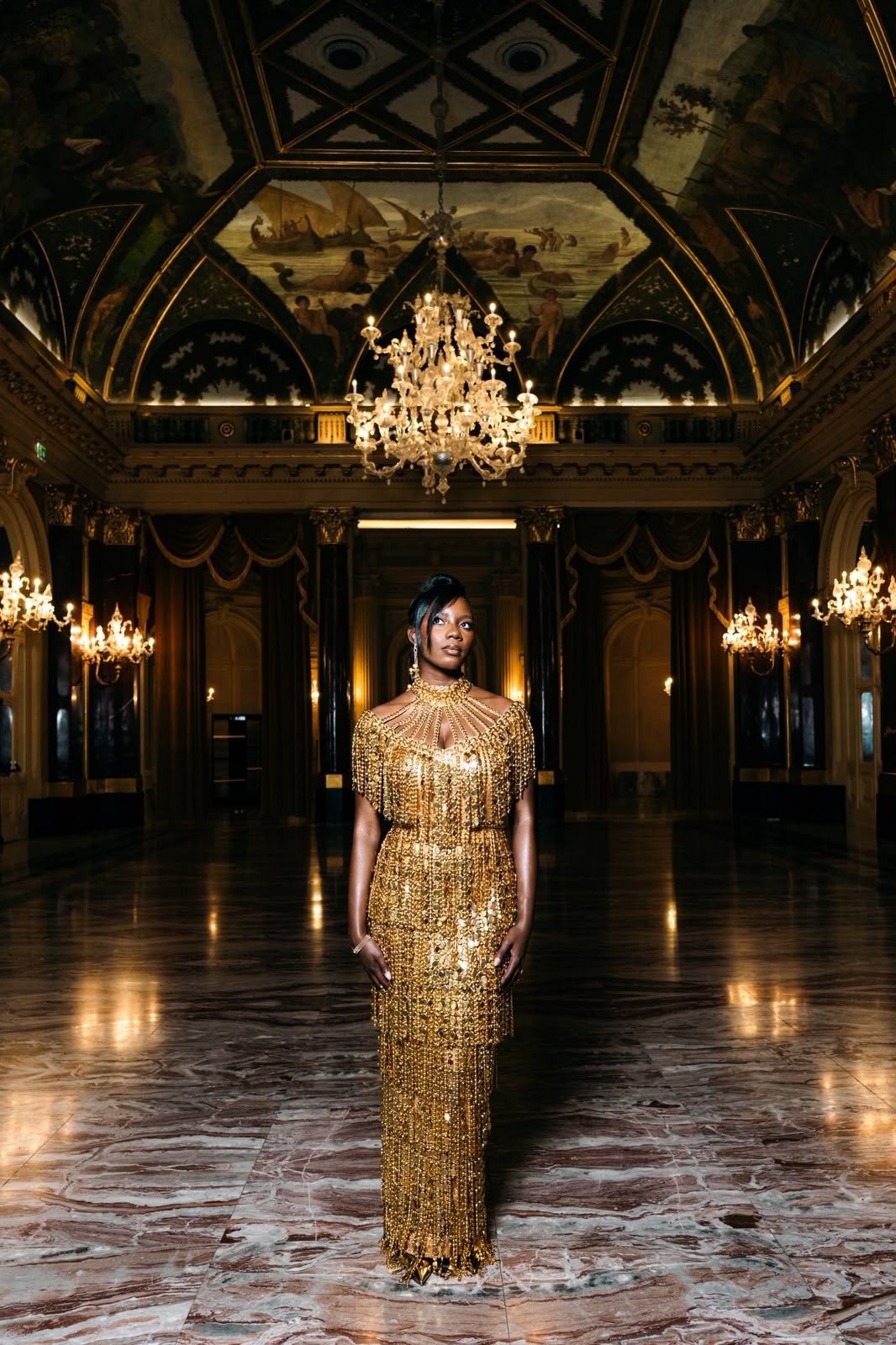 Woman in a gold beaded gown standing beneath a crystal chandelier in a dark frescoed ballroom.