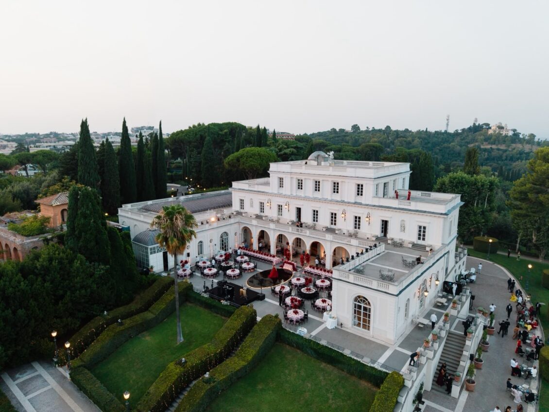 Aerial view of a white Roman villa with round dinner tables and red decor arranged on the terraces.
