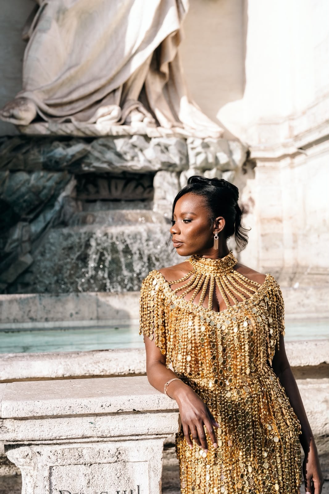 Woman in a gold beaded dress leaning beside a pale stone fountain and monumental marble sculpture.