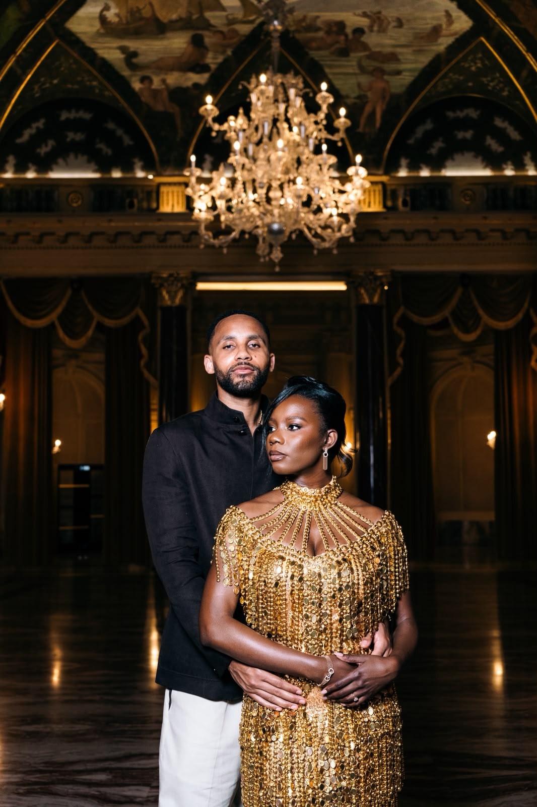 Couple posed beneath a chandelier in a dark ballroom, with the man standing behind the woman in a gold gown.