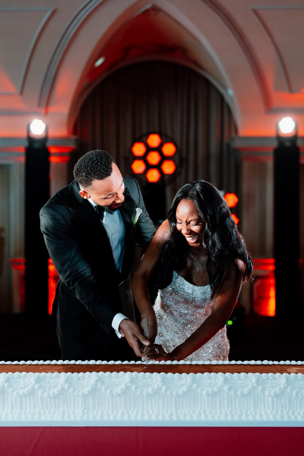 Bride and groom laughing together as they cut a long white wedding cake under red reception lighting.