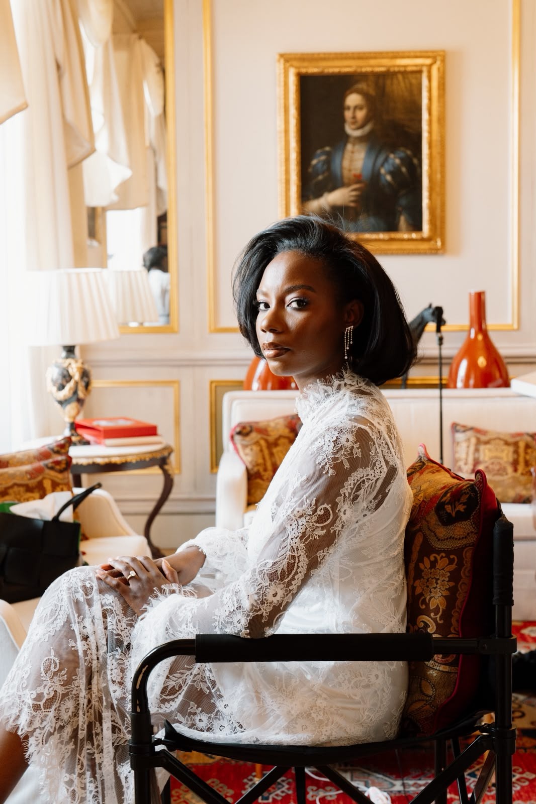 Bride seated in a patterned chair wearing a white lace robe in an elegant room with framed art.