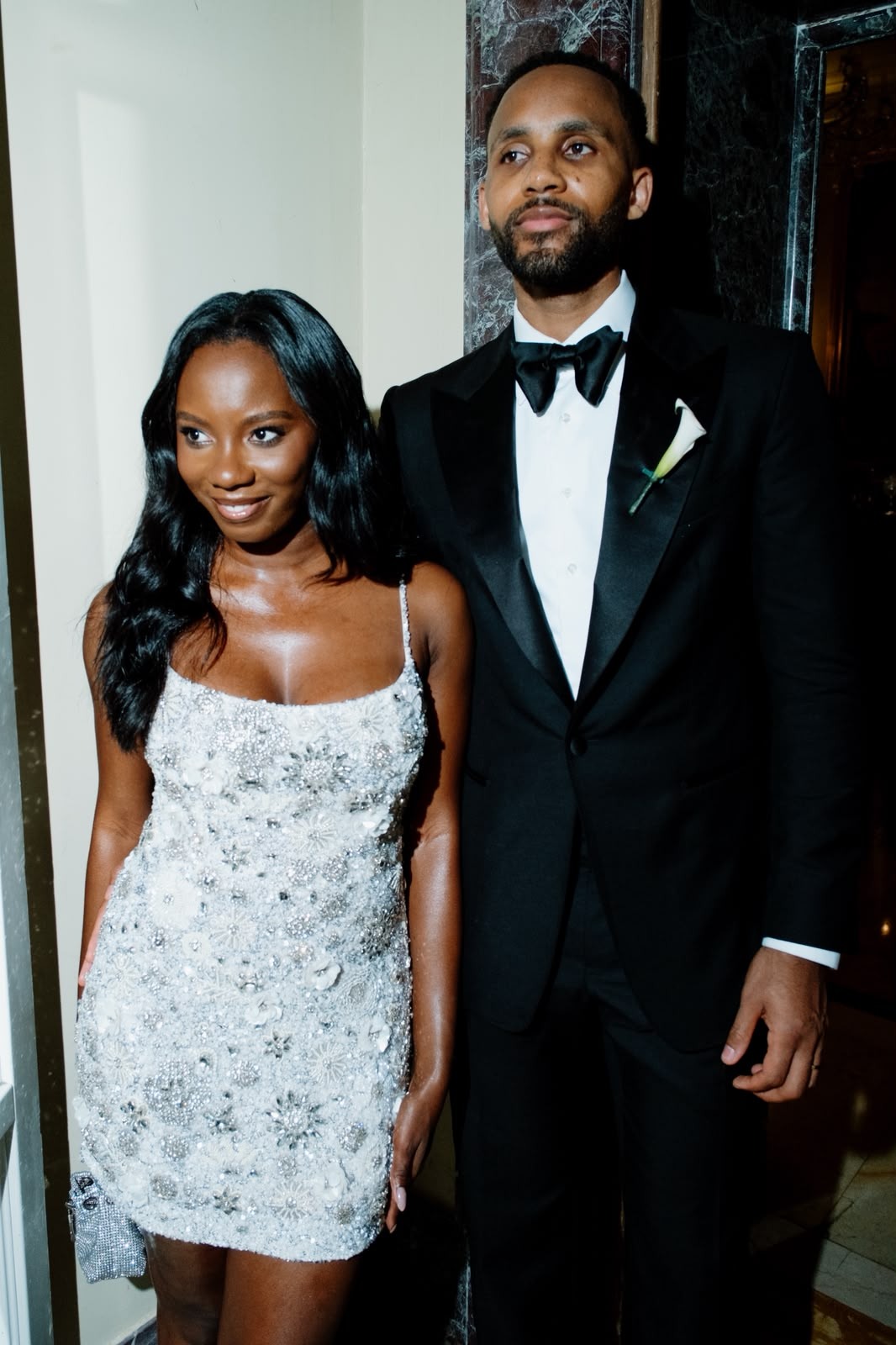 Couple posing together by a dark marble doorway, with the woman in a silver beaded dress and the man in a tuxedo.