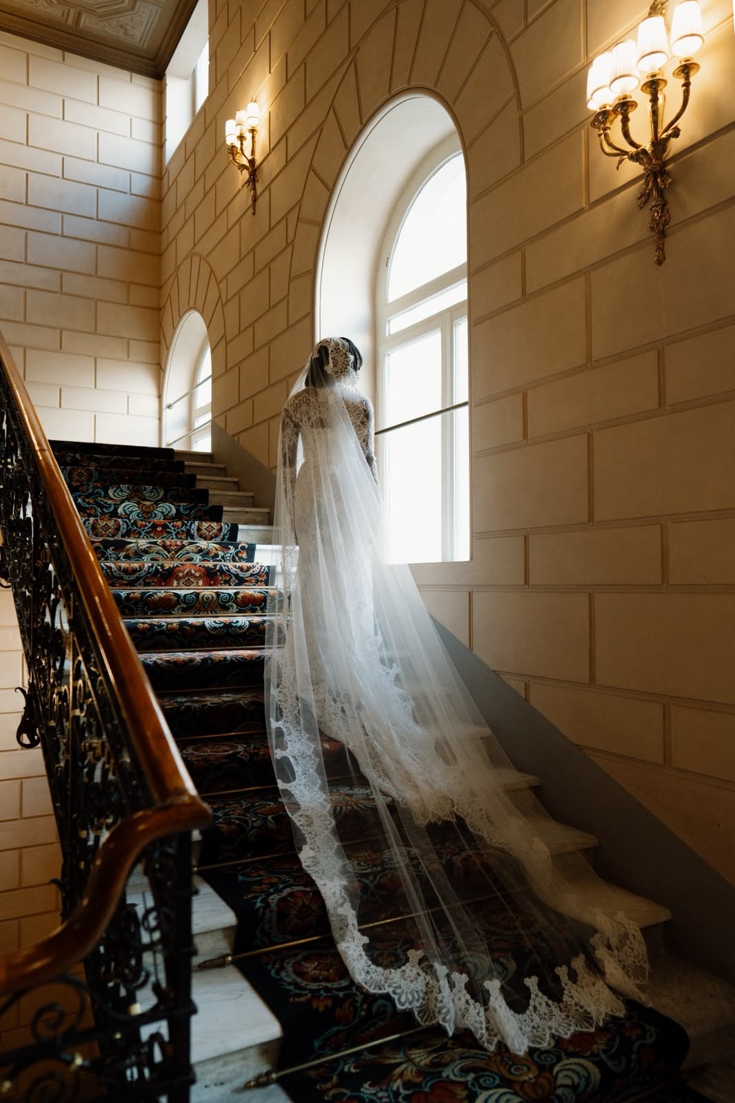 Bride in a wedding gown standing on a grand staircase with her long lace-edged veil trailing down the steps.