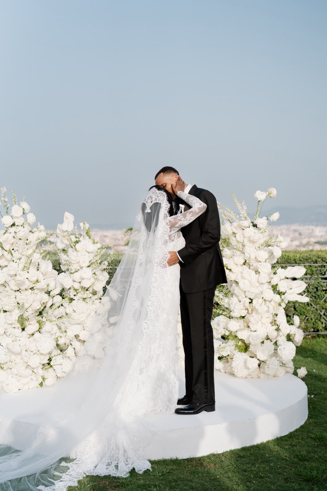 Bride in a long-sleeved lace gown and groom in a black suit kiss between white flowers at an outdoor Rome wedding ceremony.