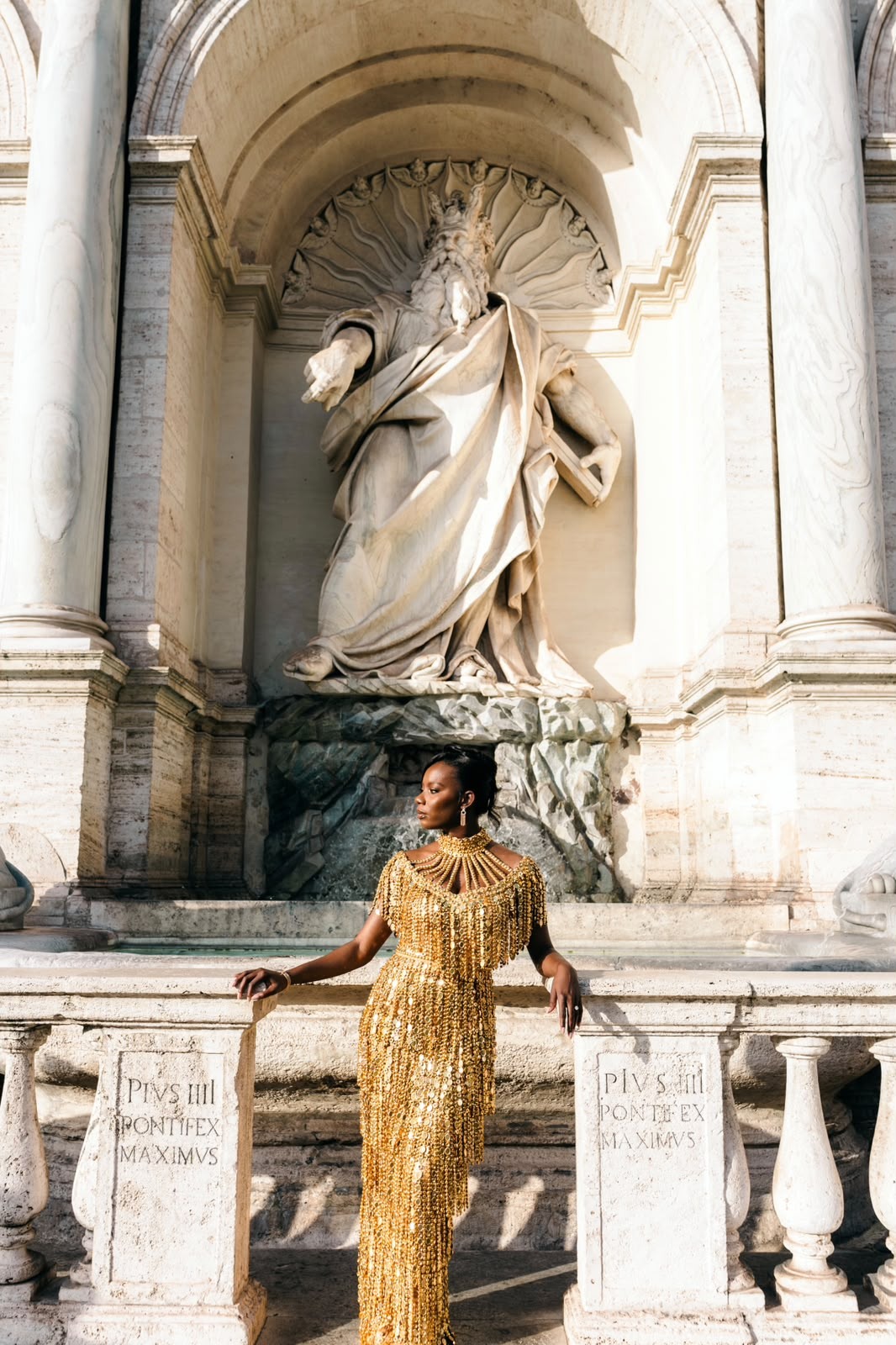 Woman in a gold beaded evening gown poses at a stone balustrade beneath a large classical statue in Rome.
