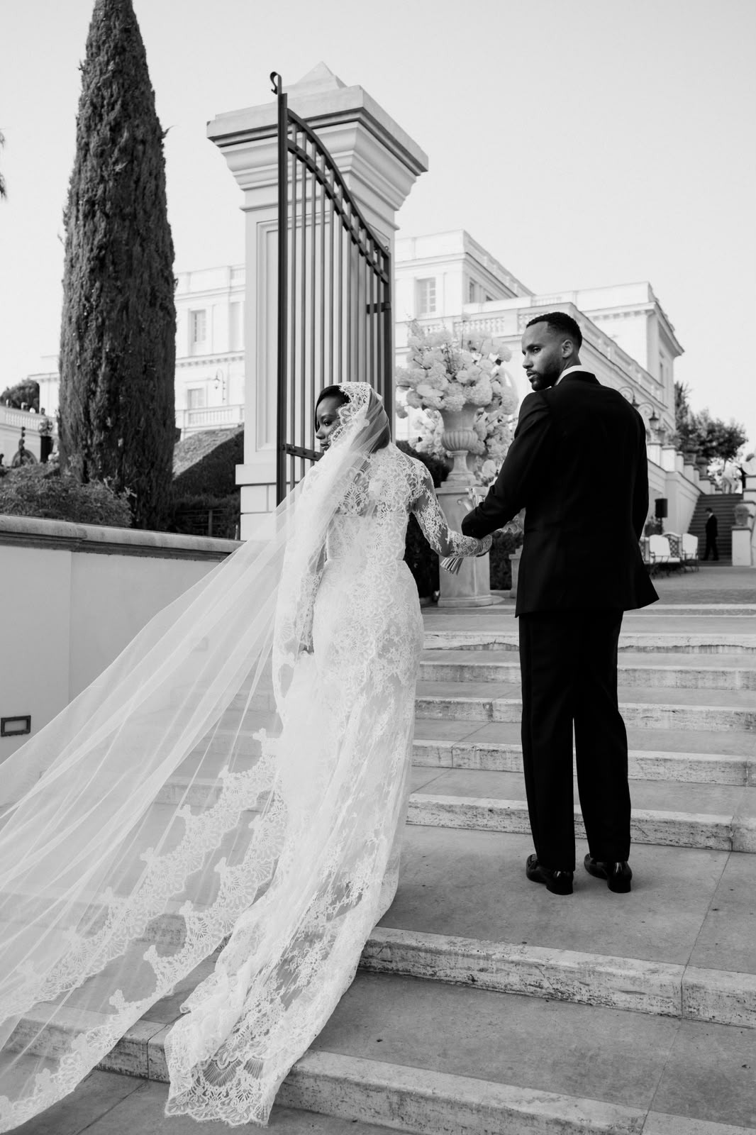 Bride and groom hold hands and look back on stone villa steps as the bride's long lace veil trails across the foreground in a black-and-white portrait.