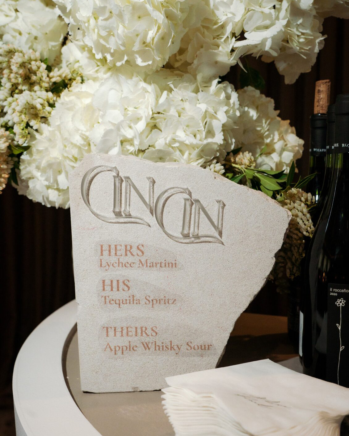 A stone drink menu with carved text reads: “CIN CIN. Hers: Lychee Martini. His: Tequila Spritz. Theirs: Apple Whisky Sour.” White flowers and wine bottles are in the background, setting a romantic scene at the Ariana and Julian Borghese wedding.
