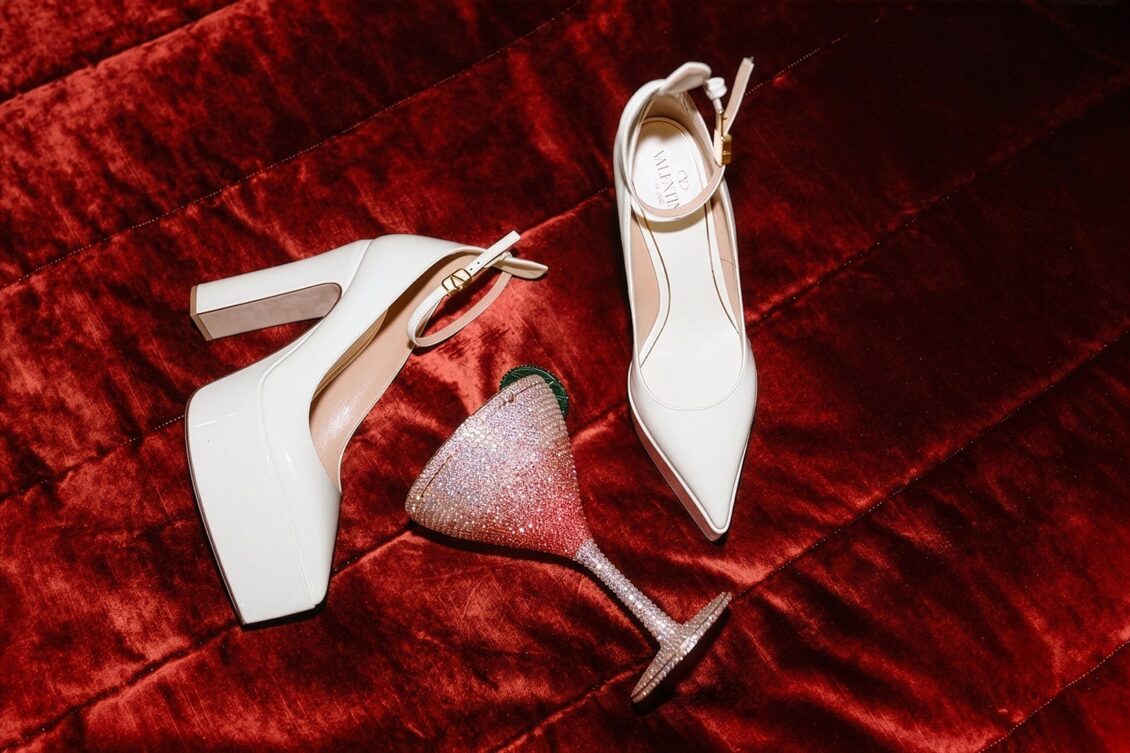 Two white high-heeled shoes and a glittery, martini glass-shaped clutch lie on a rich red velvet surface, capturing the elegant details of an Irish castle wedding at Ashford Castle.